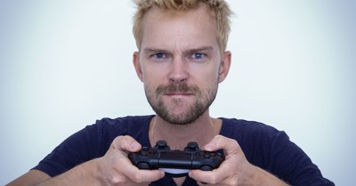 Man with short blonde hair fiercely focused while gaming with a controller against white background.