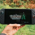 A hand holding a handheld gaming console displaying the Pokémon Legends game screen outdoors.